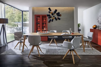 Großer moderner Esstisch aus hellem Holz, um den sechs Stühle mit grauem Stoffbezug stehen mit und ohne Armlehne und hellem Holzgestell.